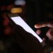 Close-up of a hand using a smartphone in the dark with blurred city lights in the background.