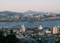 Stunning view of a bustling port city at sunrise with cranes, buildings, and mountains.