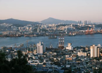 Stunning view of a bustling port city at sunrise with cranes, buildings, and mountains.