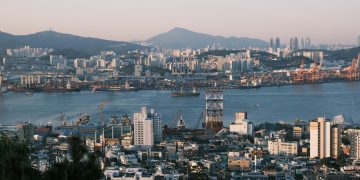 Stunning view of a bustling port city at sunrise with cranes, buildings, and mountains.