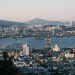 Stunning view of a bustling port city at sunrise with cranes, buildings, and mountains.