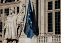 European Union flag waving beside a historic statue on stone building facade.