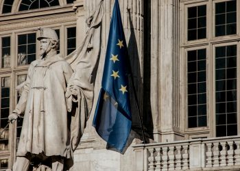 European Union flag waving beside a historic statue on stone building facade.