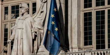 European Union flag waving beside a historic statue on stone building facade.