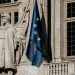 European Union flag waving beside a historic statue on stone building facade.