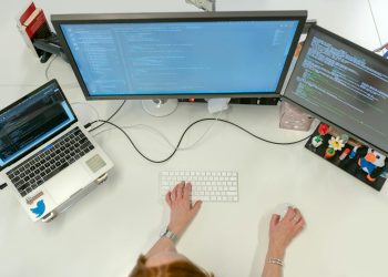 A female software engineer coding on dual monitors and a laptop in an office setting.