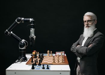 A robotic arm and a bearded man engaged in a strategic chess game highlighting technology and innovation.