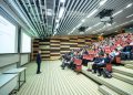 A speaker delivers a presentation to an attentive audience in a modern auditorium setting.