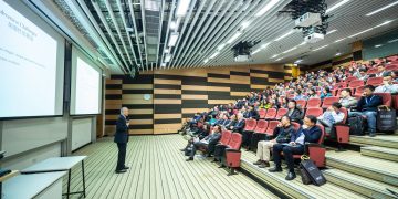 A speaker delivers a presentation to an attentive audience in a modern auditorium setting.