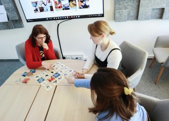 Three women playing a card game at an office table, fostering teamwork.