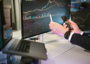 A professional analyzing financial charts on computer monitors and a smartphone, symbolizing digital finance strategies.