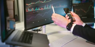 A professional analyzing financial charts on computer monitors and a smartphone, symbolizing digital finance strategies.