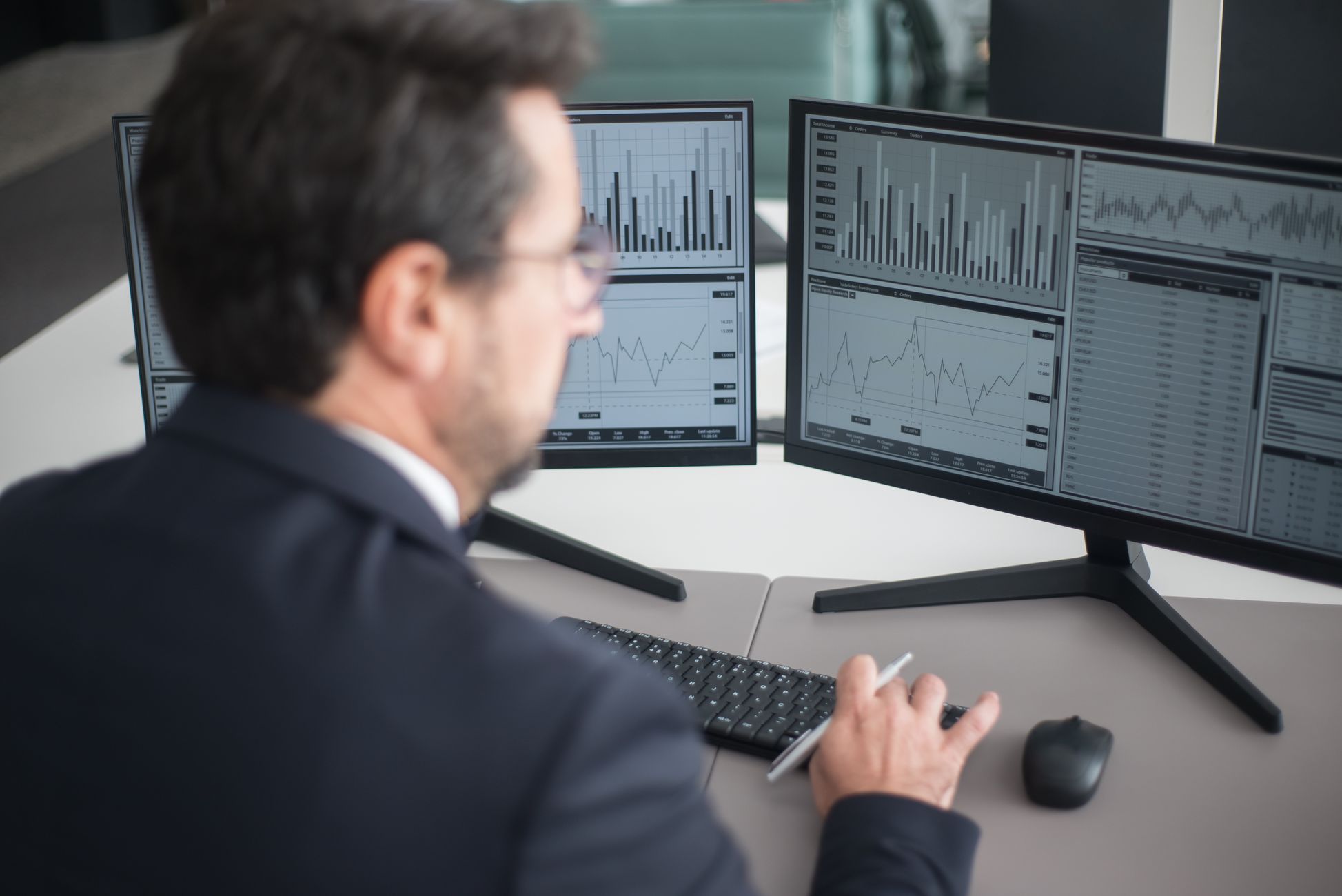 A businessman in a suit analyzing financial charts and graphs on two computer monitors indoors.