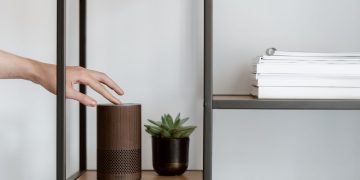 A hand interacts with a smart speaker on a minimalist shelf, featuring a plant and stacked books.