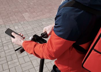 A delivery person riding an electric scooter using a smartphone for navigation outdoors.