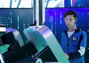 Teenager interacts with futuristic device using VR goggles in tech environment.