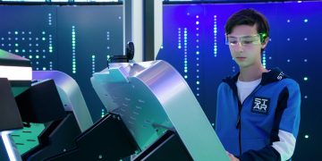 Teenager interacts with futuristic device using VR goggles in tech environment.
