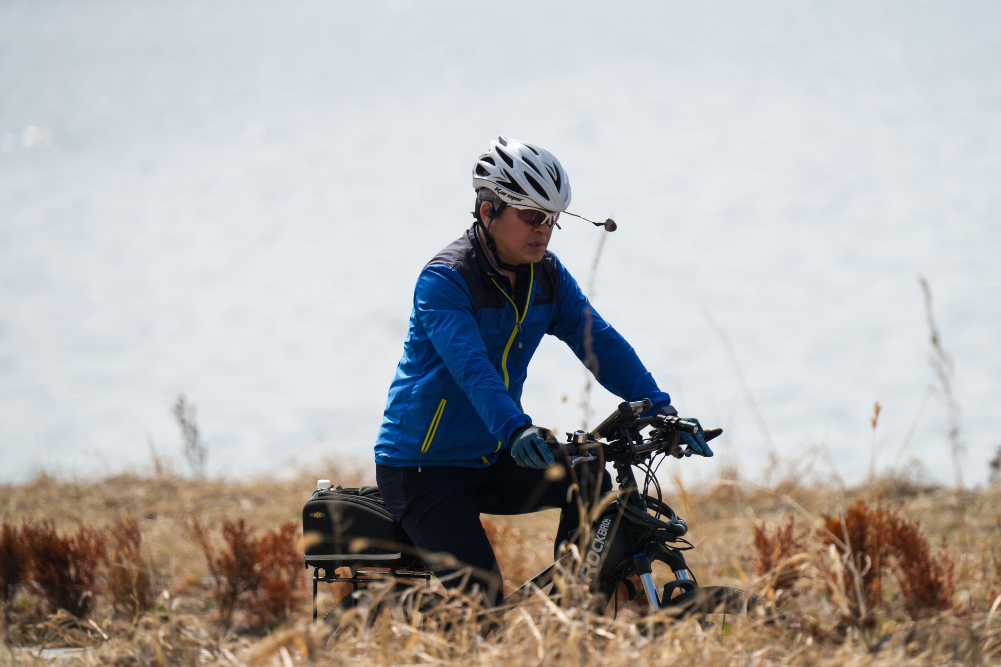 Adult cyclist in blue jacket riding by a lake with dry grass foreground, wearing helmet and sunglasses.