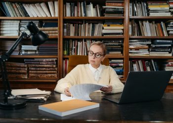 A senior woman focuses on reading documents at a desk in a library, surrounded by books and a laptop.