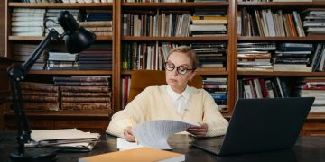 A senior woman focuses on reading documents at a desk in a library, surrounded by books and a laptop.