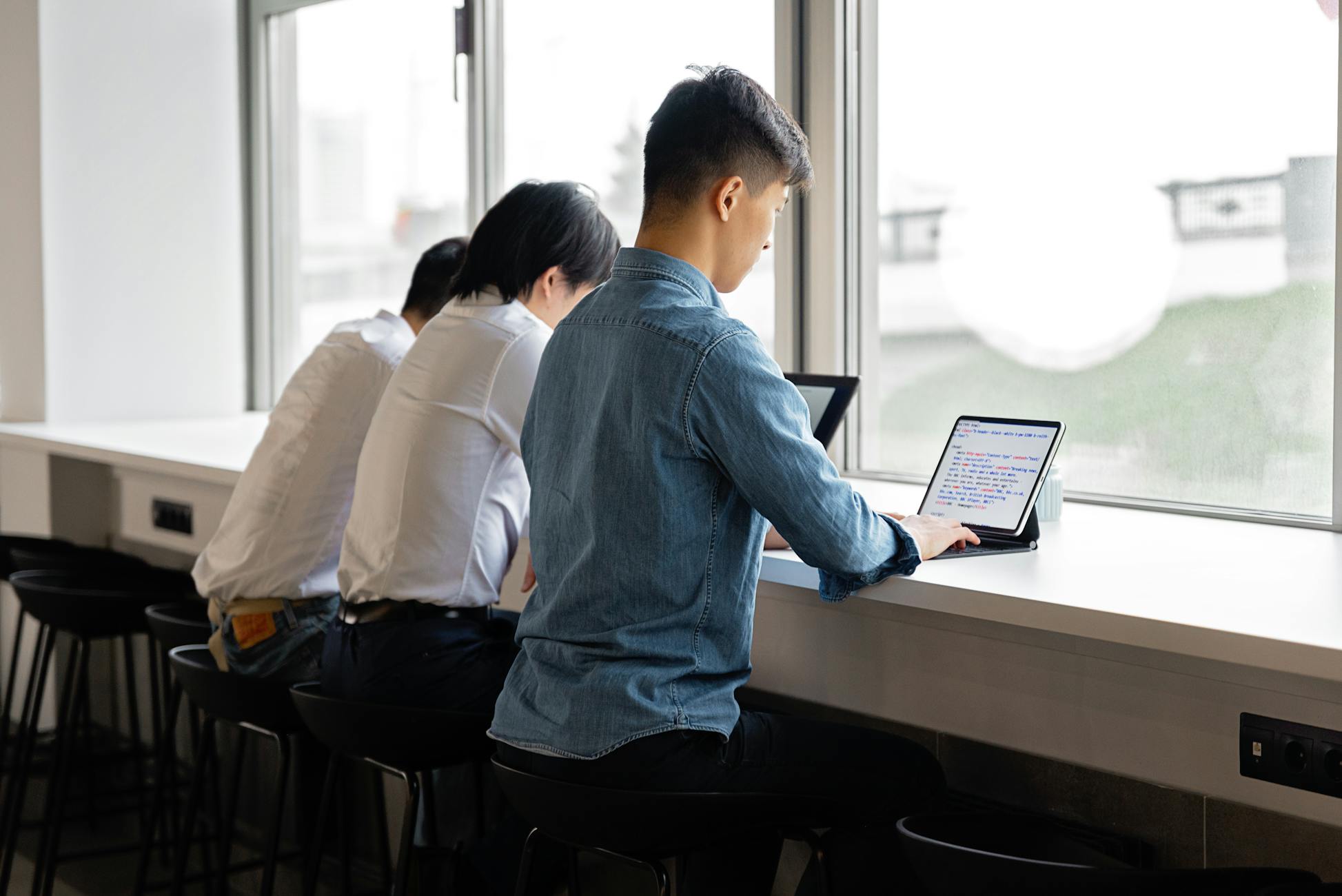 A group of young adults sitting by a window using laptops in a modern office interior.