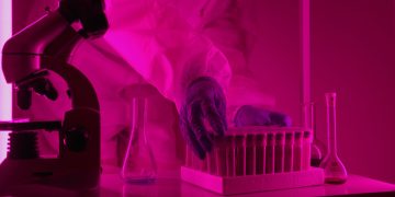 Scientist in a lab coat handling test tubes under pink lighting, using a microscope.