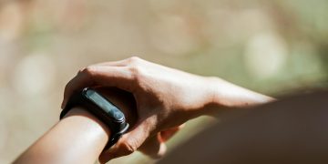 From above of crop anonymous person wearing modern bracelet with small display while standing outdoors on blurred background in daylight