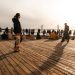 A group of people enjoying a sunny day on a wooden dock with a bridge in the background.