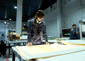 Worker in uniform examining sheets in a brightly lit factory environment, focusing on quality.
