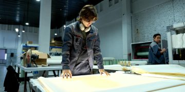 Worker in uniform examining sheets in a brightly lit factory environment, focusing on quality.