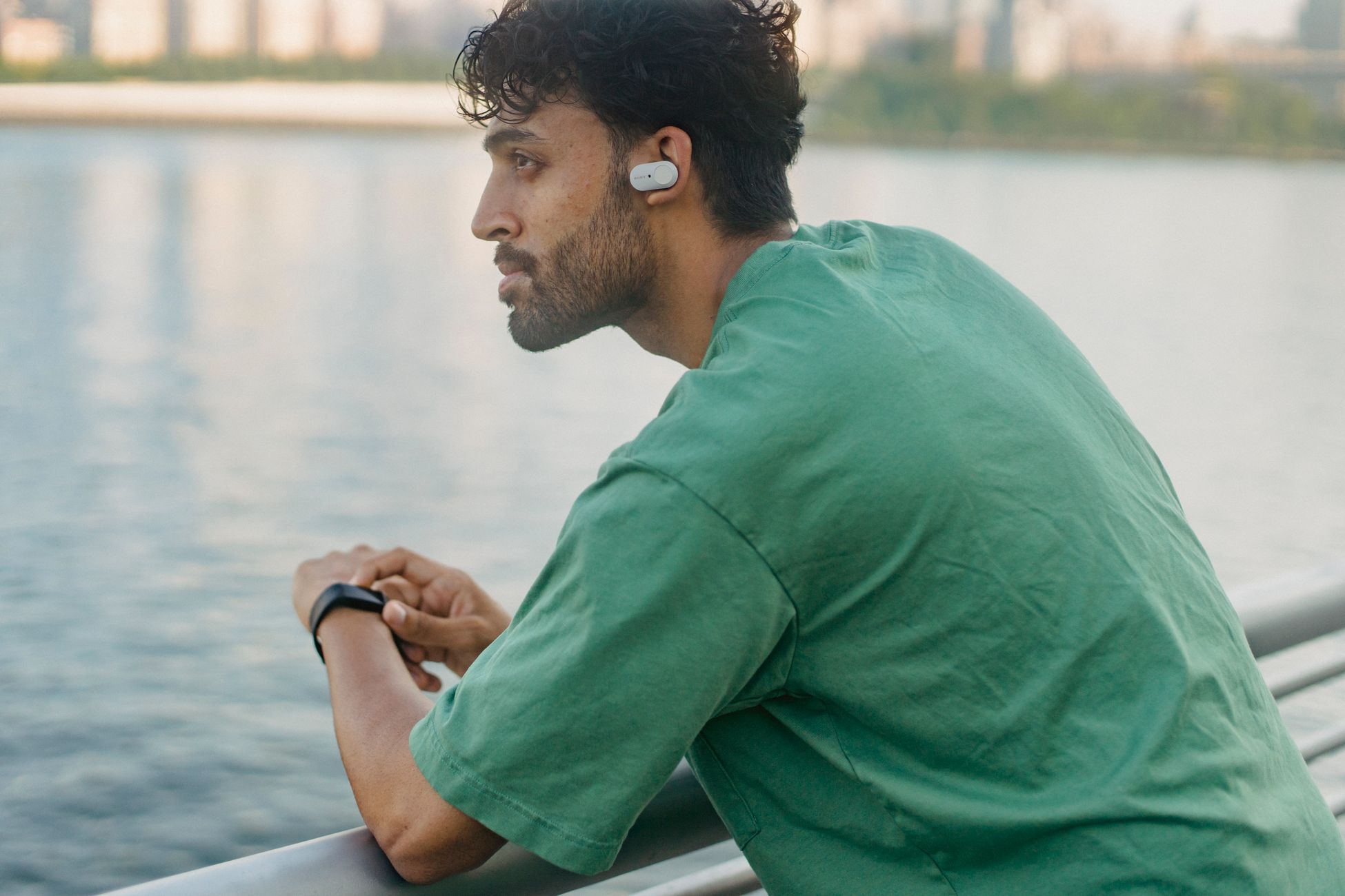 Contemplative man by waterfront, wearing a green shirt and earpiece, leaning on railing.