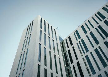 From below of contemporary multistory building with futuristic design and windows with unusual shapes located against cloudless blue sky on street of city