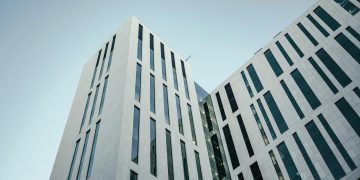 From below of contemporary multistory building with futuristic design and windows with unusual shapes located against cloudless blue sky on street of city