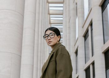 Portrait of a stylish woman in eyeglasses, olive coat, against modern architectural backdrop.