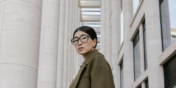 Portrait of a stylish woman in eyeglasses, olive coat, against modern architectural backdrop.