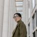 Portrait of a stylish woman in eyeglasses, olive coat, against modern architectural backdrop.
