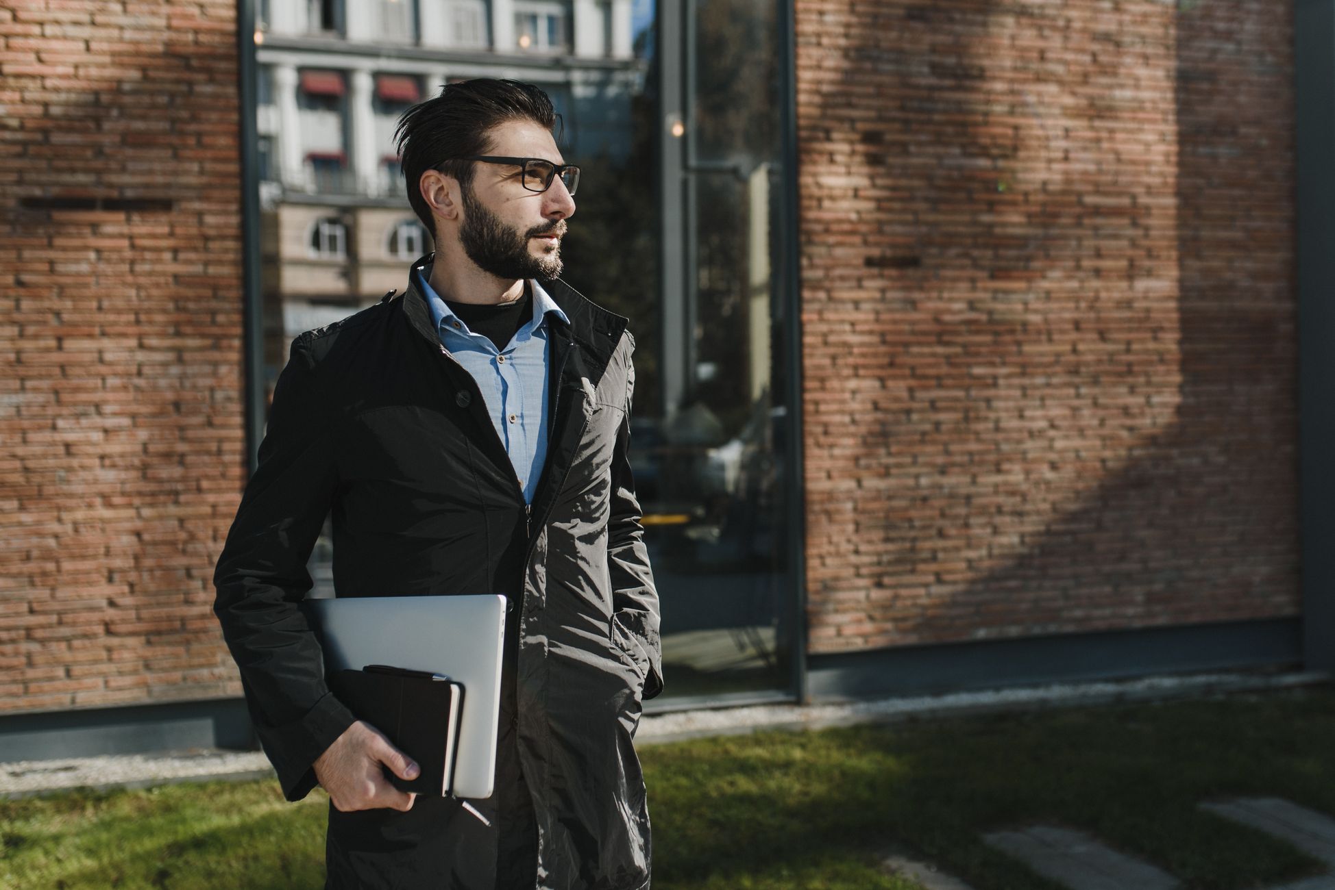 Stylish man in glasses with a laptop standing outdoors against a brick wall, looking confident.