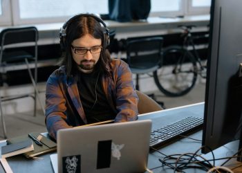 Bearded male developer in headphones coding on a laptop in a modern office setting.