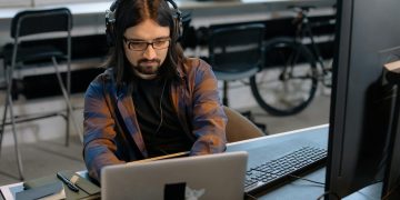 Bearded male developer in headphones coding on a laptop in a modern office setting.