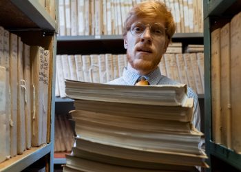 Man with glasses holding a stack of documents in a library archive, surrounded by shelves of files.