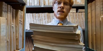 Man with glasses holding a stack of documents in a library archive, surrounded by shelves of files.