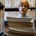 Man with glasses holding a stack of documents in a library archive, surrounded by shelves of files.