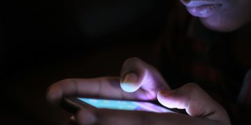 Close-up of a person holding and using an illuminated smartphone in a dark setting.
