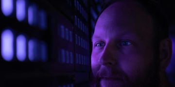 Close-up of a bearded man looking contemplatively at a purple-lit wall.