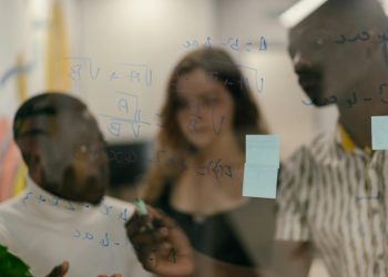 Diverse group of people solving math equations on a glass panel indoors.