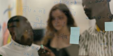Diverse group of people solving math equations on a glass panel indoors.