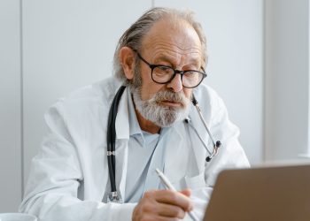 Elderly doctor with glasses and stethoscope working on a laptop in a medical office.