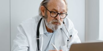 Elderly doctor with glasses and stethoscope working on a laptop in a medical office.