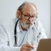 Elderly doctor with glasses and stethoscope working on a laptop in a medical office.