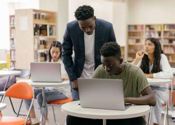 Students learning in a classroom setting with a teacher assisting and laptops on desks, creating an interactive education environment.